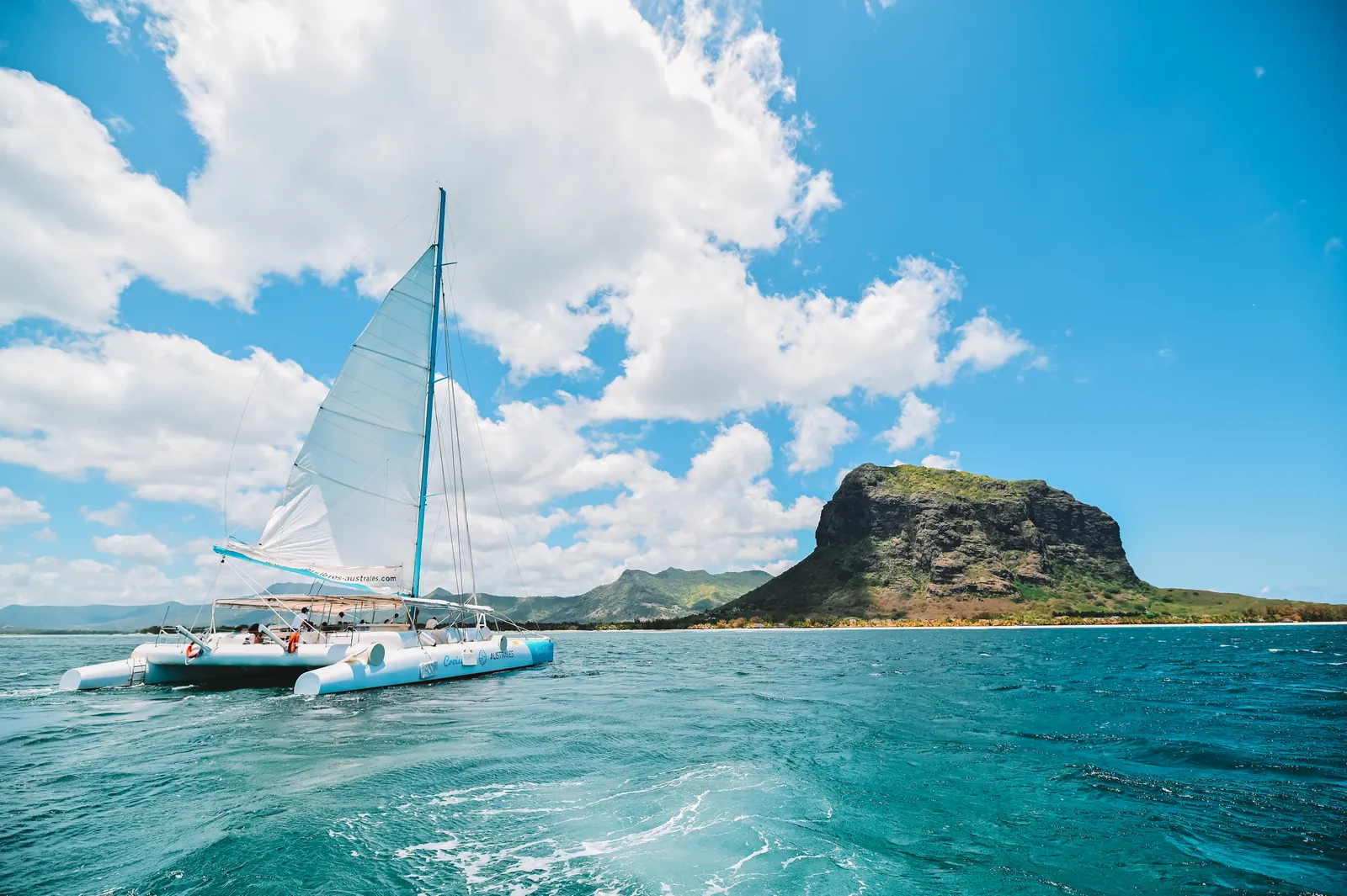 A catamaran sailing with a rocky mountain behind it.